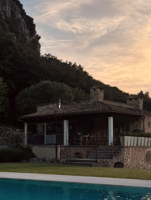 Stone house with tan brick and glass facade, lush green trees, and a rectangular pool in the foreground. Mountains and dusk sky in the background.