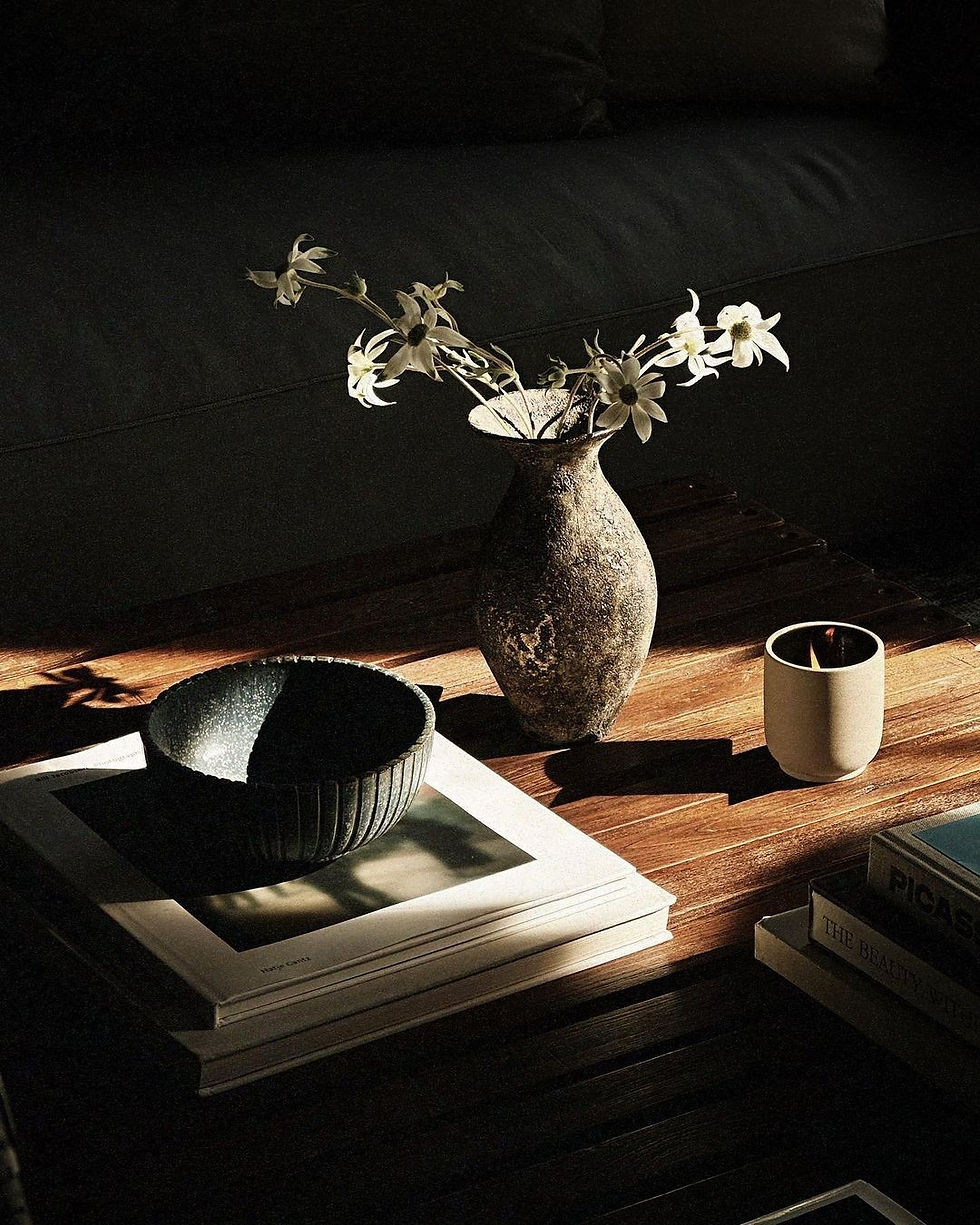 Ceramic vase with white flowers on a wooden table. Shadow play highlights a black bowl, books, and a cup in a cozy, dimly-lit room.