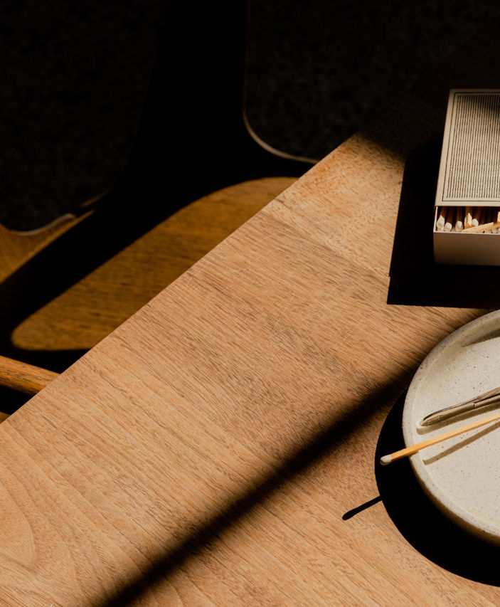 Wooden table with a box of matches and a white plate holding chopsticks. Sunlight creates shadows, adding a warm, calm atmosphere.