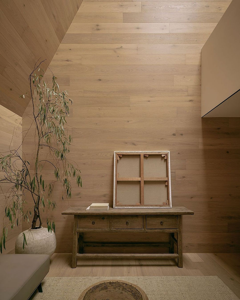 Minimalist room with wooden walls, a large vase with a leafy plant, a rustic table holding books, and an empty canvas frame leaning against the wall.