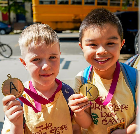 Two boys in yellow Park Slope Day Camp shirts holding medals out to the camera-- the left with a letter A, the right with a letter K
