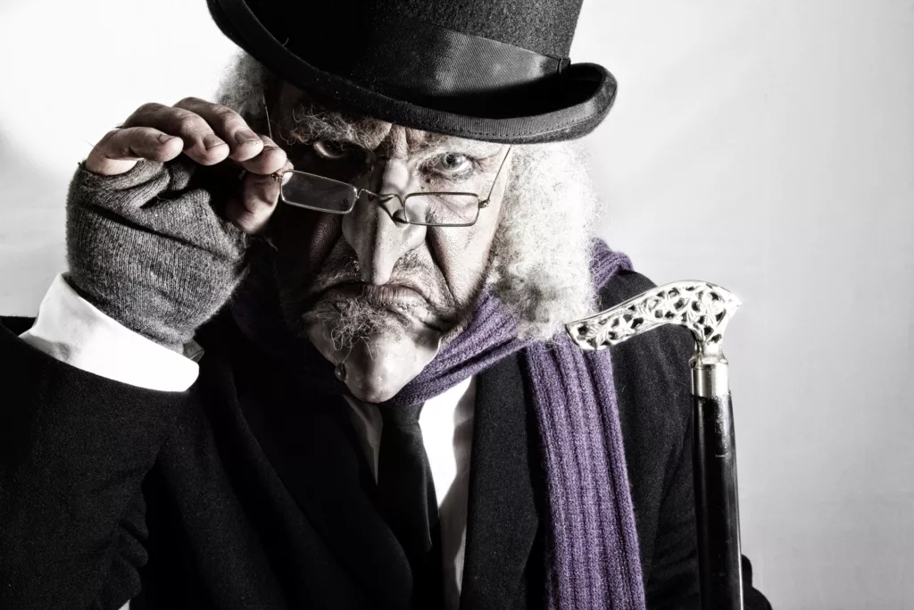 stern looking mature man in victorian costume carrying a cane  . Model is wearing a dark suit , top hat, glasses and prosthetic make up , the look created is also similar to a  Dickens victorian type character .