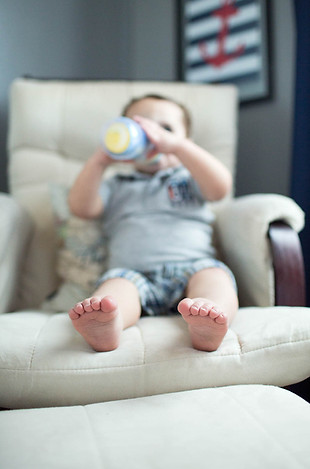 toddler drinks from his bottle sitting in his nursery rocking chair in their Herriman City, Utah Home