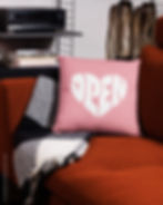 Light pink pillow with a white logo that says “open,” with the letters forming the shape of a heart, on a sofa