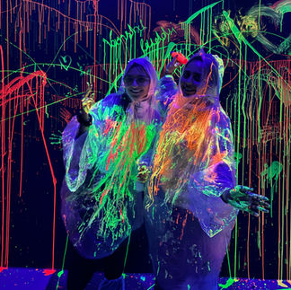Two people wearing transparent ponchos showing their neon-painted hands under blacklight in a glow-in-the-dark paint room with colorful splashes on the walls and floor.