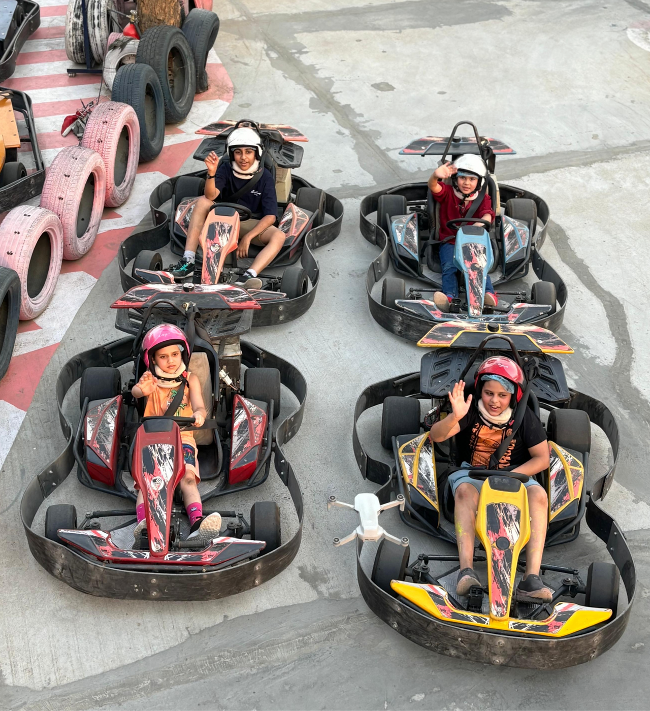 Four people in go-karts lined up in rows, smiling and waving while wearing helmets at an outdoor go-kart track.