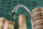 Torneira de água com moedas ao fundo. Entenda os motivos do aumento nas tarifas de água em todo o mundo