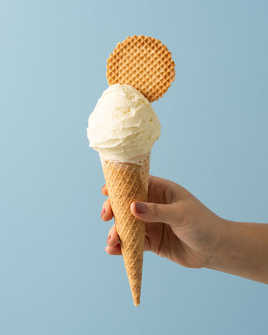 A scoop of vanilla gelato in a waffle cone with a waffle disc is being held in front of a blue background.