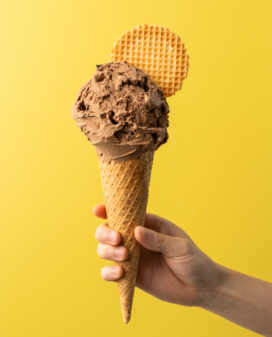 An image of a scoop of chocolate gelato in a waffle cone and a waffle disc held in front of a yellow background
