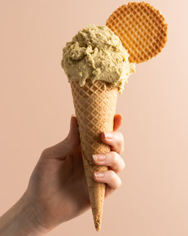 A scoop of pistachio gelato in a waffle cone with a waffle disc is being held in front of a light pink background.