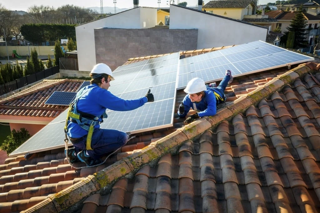 Un homme accroupis et une femme allongé, portant tout deux des équipement de protection individuelles mais sans aucune attache. Il installent un panneau solaire