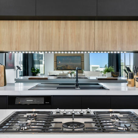 Modern kitchen with black and wood cabinets, a gas stove, and a sink. A mirror reflects a bright living room. Minimalistic decor.