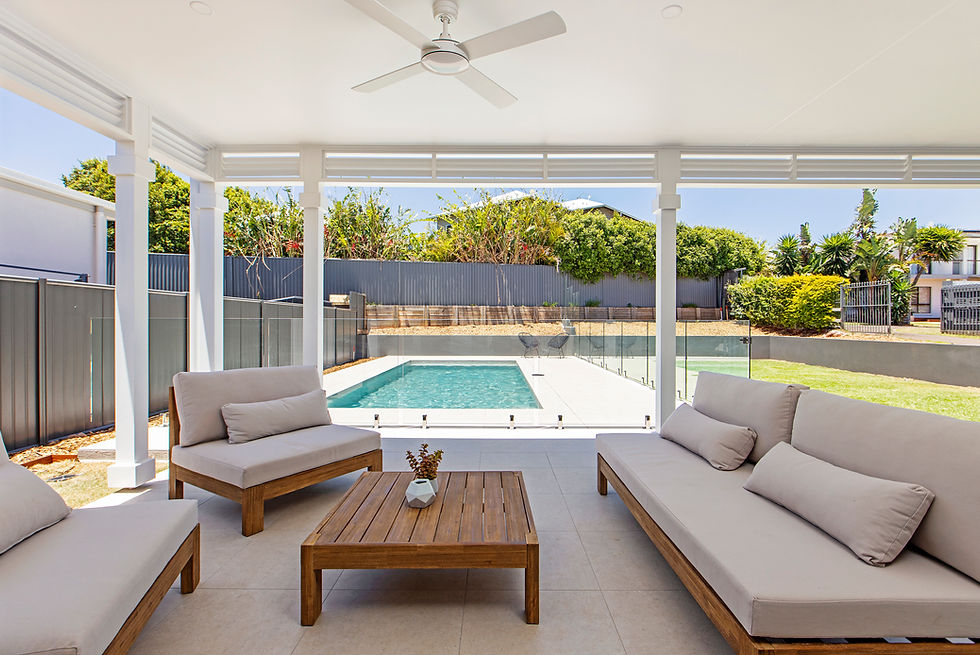 Outdoor patio with beige sofas, a wooden table, and a ceiling fan overlooking a clear blue pool and green garden on a sunny day.