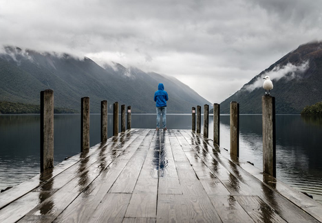 Person in blue jacket standing on pier overlooking mountains and lake.