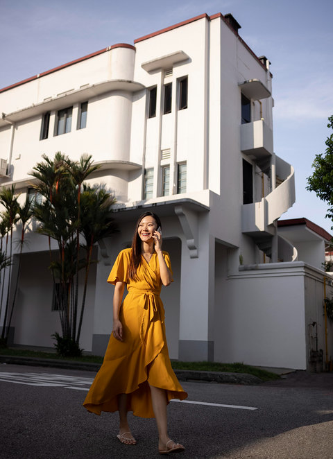 Woman in yellow dress talking on phone near a white building.