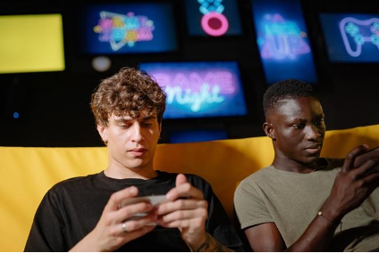 Two men focused on their phones sit on a yellow couch with neon signs in the background. Mood is concentrated and relaxed.