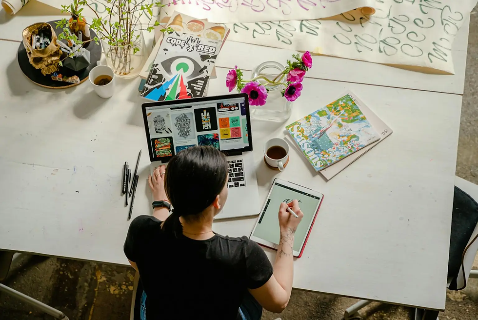 Person drawing on a tablet at a desk with a laptop displaying designs, surrounded by art supplies, magazines, and a coffee cup. Bright, creative scene.