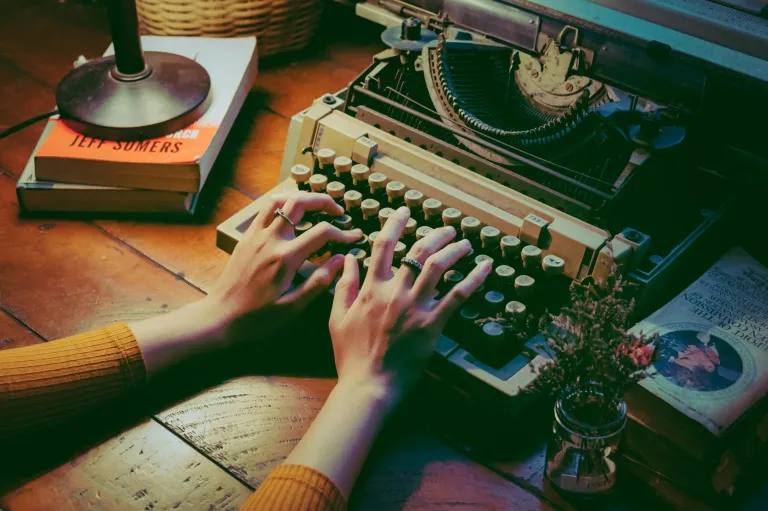 Hands typing on a vintage typewriter on a wooden table with books, a lamp, and flowers. Warm lighting creates a nostalgic ambiance.