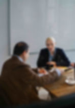 Two people in business attire sit at a wooden table with coffee cups and documents, engaged in conversation. A whiteboard is in the background.