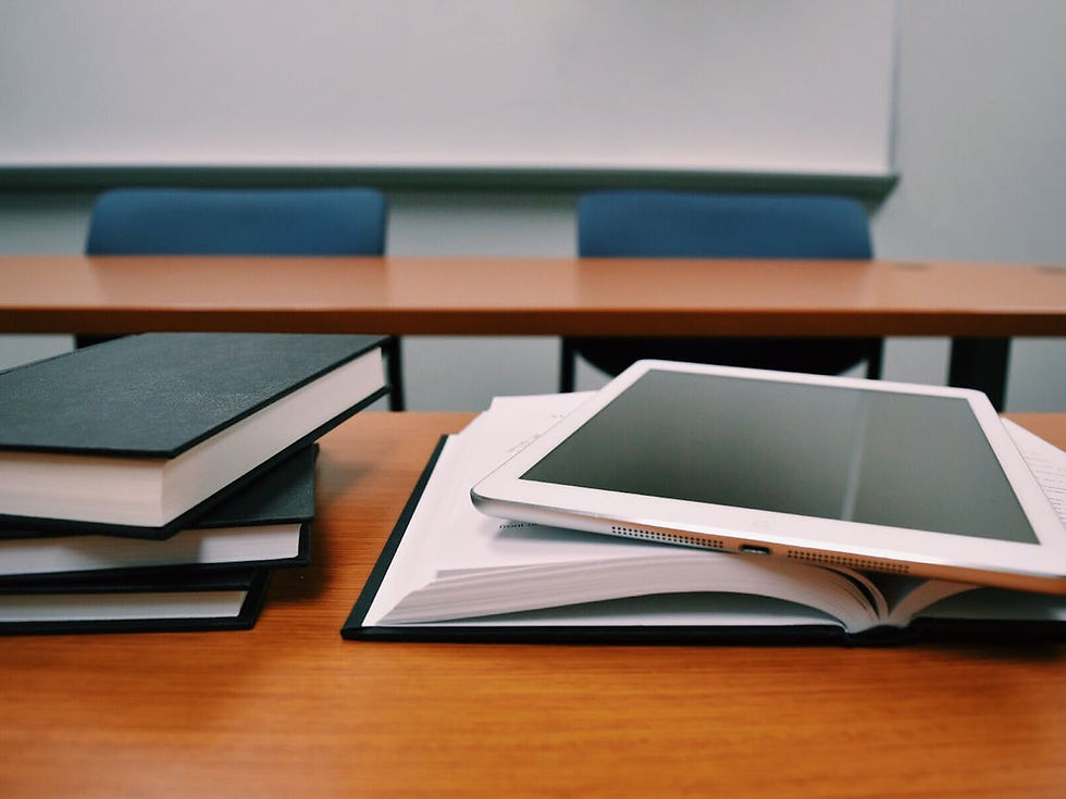Tablet on a book, stacked books nearby on a wooden desk in a classroom. Blue chairs and beige board in background. Calm academic setting.