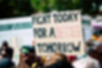 People at a protest, holding a sign that reads, "FIGHT TODAY FOR A BETTER TOMORROW," with a blurred background showing trees and a truck.