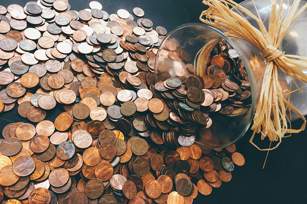 Coins spilling from a glass jar tied with straw, scattered over a dark surface. Predominantly bronze and copper tones.