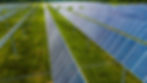 Solar panels arranged in rows on green grass, capturing sunlight. Clear blue sky and forested background. Bright, efficient, renewable energy.