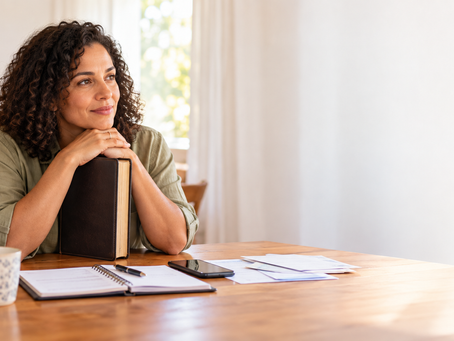 Adulto em ambiente doméstico contemporâneo observa calmamente vários objetos de uso diário sobre uma mesa enquanto segura uma Bíblia fechada, em cena sóbria de reflexão.