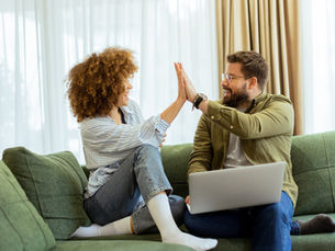 Man and woman high-five on green sofa. Man holds laptop; both smiling. Bright room with curtains; casual, happy mood.