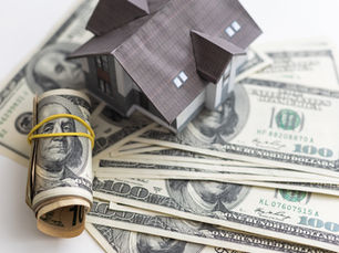 Miniature house on stacked $100 bills and rolled cash tied with a yellow band. Represents real estate finance, white background.