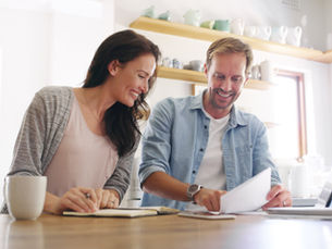 Smiling couple reviews documents at a wooden table with a laptop, notebook, and mug in a bright kitchen. Shelves hold pastel ceramics.