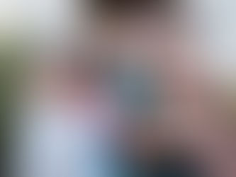 Smiling family of four outdoors. A man holds a child in a brown dress; a woman embraces another child in pink, with greenery in the background.