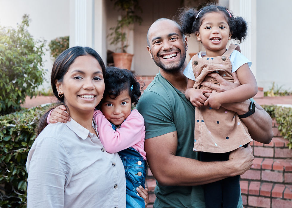 Smiling family of four outdoors. A man holds a child in a brown dress; a woman embraces another child in pink, with greenery in the background.