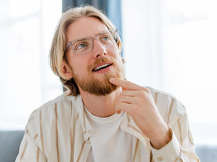 Man with glasses and a beard, thoughtfully touching chin, wearing a striped shirt. Bright, blurred interior background, conveying curiosity.