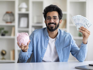 Smiling man holds pink piggy bank and fan of cash in office with white shelves. Wears blue shirt and glasses, expressing happiness.