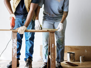 Two people in gloves cut wood on a sawhorse using a red saw. Paint-splattered jeans; paint cans and a brush visible on the floor.