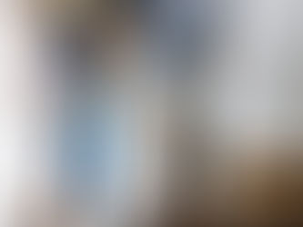 Two people in gloves cut wood on a sawhorse using a red saw. Paint-splattered jeans; paint cans and a brush visible on the floor.