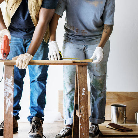 Two people in gloves cut wood on a sawhorse using a red saw. Paint-splattered jeans; paint cans and a brush visible on the floor.