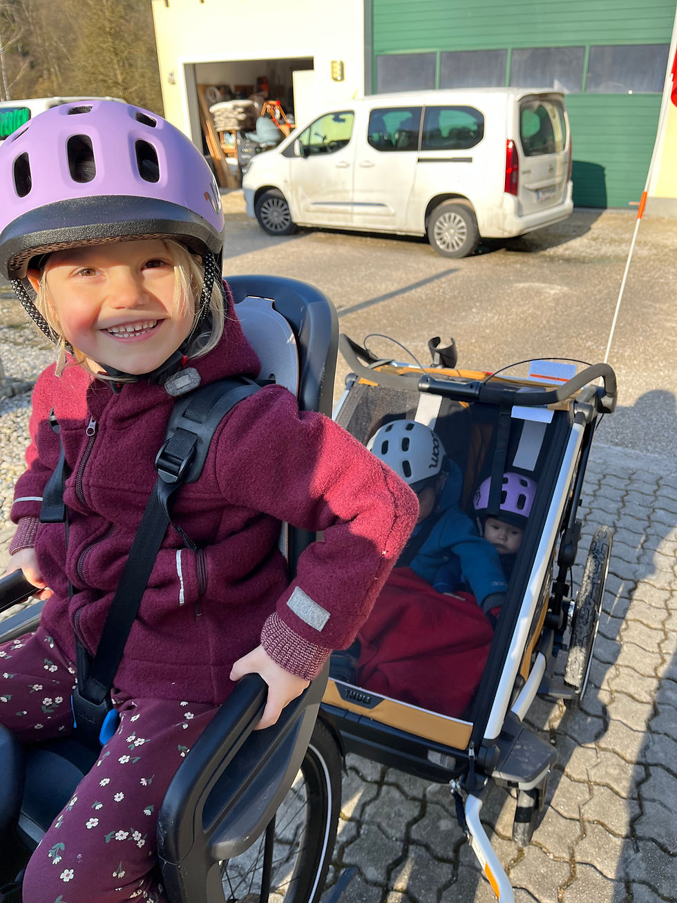 2 Kinder im Fahrradanhänger Thule Chariot Sport2 Double mit Helmen von Woom und 1 Kind am Fahrradsitz von Thule dem RideAlong2