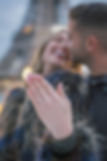 Paris proposal engagement photoshoot A romantic moment captured in a Parisian street as a man kneels down to propose to his partner, who looks surprised and emotional. The couple is surrounded by colorful buildings and trees, and the Eiffel Tower can be seen in the distance. A photographer stands nearby, capturing the special moment on camera.