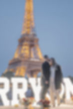 Couple embracing in front of lit Eiffel Tower, with "MARRY" sign, candles, and flowers around. Romantic evening setting.