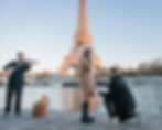 A couple in Paris, with the Eiffel Tower visible in the background. The man is down on one knee, holding out a ring to the woman, who has her hands covering her face in surprise and excitement. A photographer captures the moment, with the Seine River and a row of trees in the background.