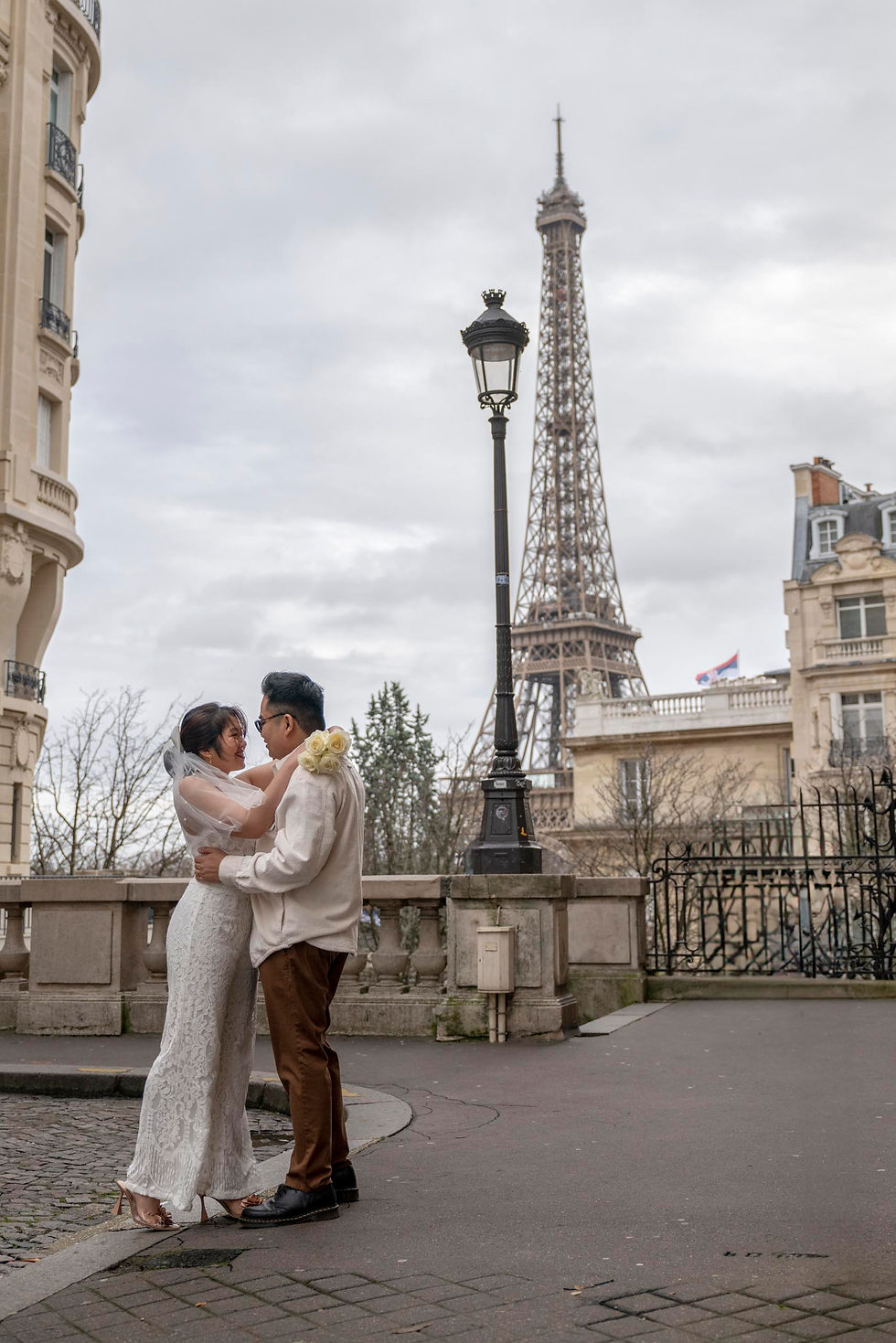 Couple embraces near the Eiffel Tower on a cloudy day. She wears a white dress and veil, holding yellow roses. Parisian buildings surround them.