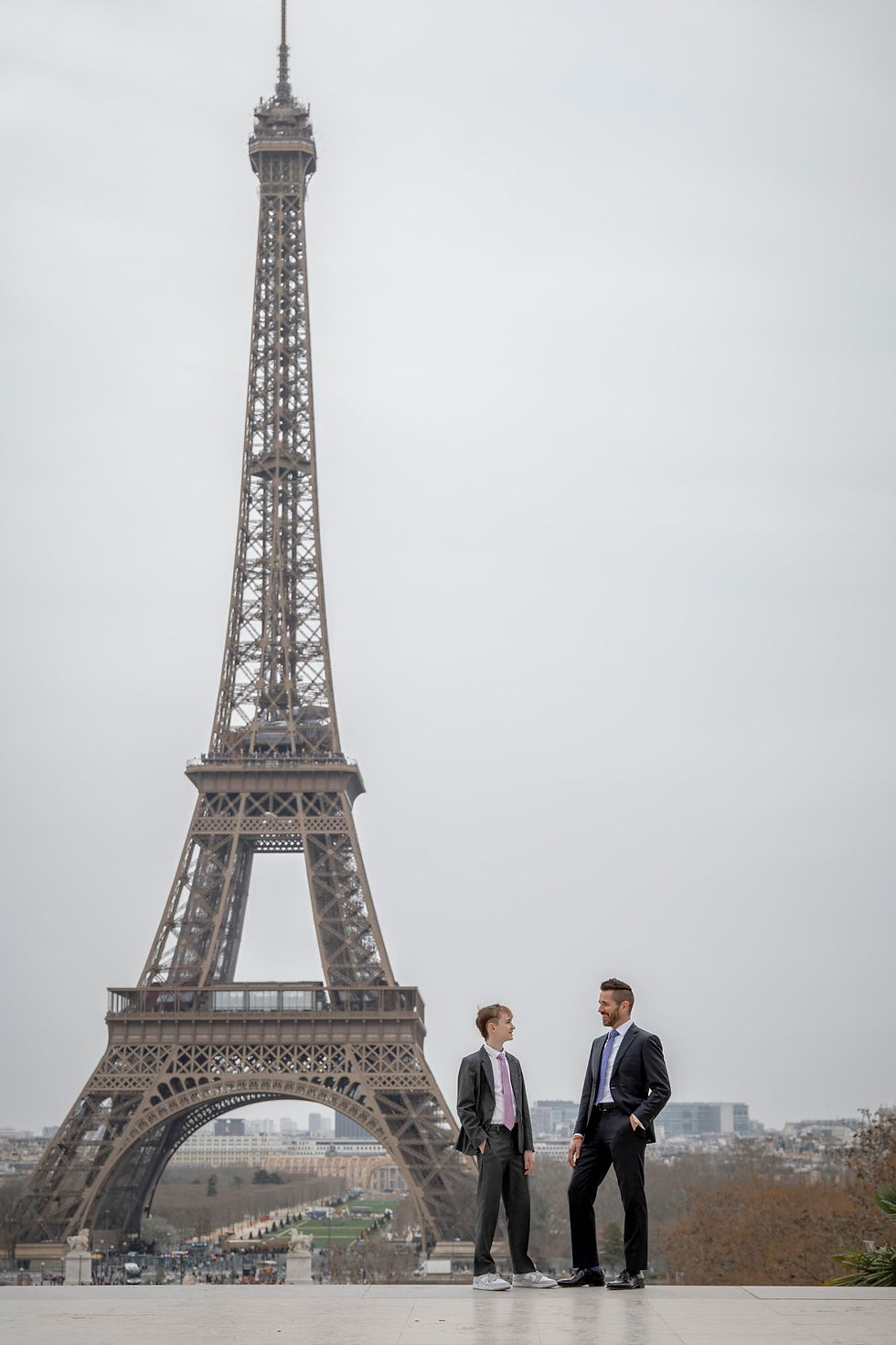 Two people in suits stand conversing in front of the Eiffel Tower on a cloudy day. The mood is casual and relaxed. No text visible.