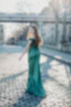 Woman in a green gown walks on a cobblestone street, looking back with a smile. Sunlight filters through trees and a bridge in the background.