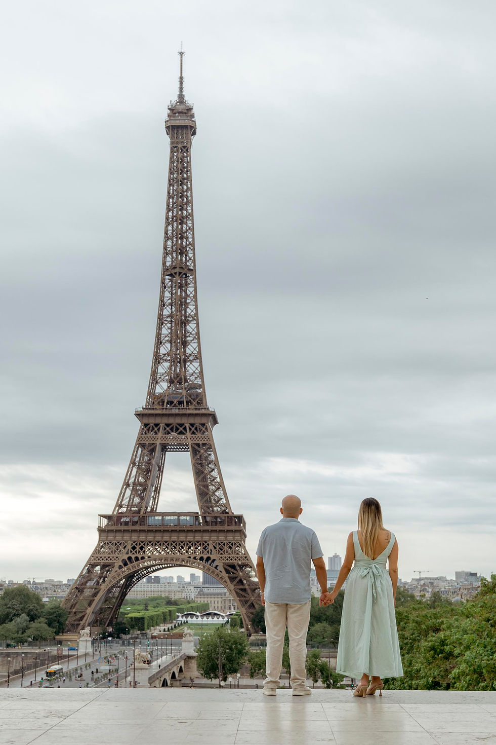 A couple holding hands faces the Eiffel Tower on a cloudy day. The woman wears a light green dress. Paris cityscape in the background.