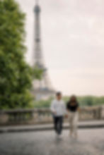 A couple walks hand in hand on a paved street with the Eiffel Tower in the background. They are smiling, surrounded by greenery.