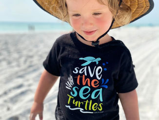 A toddler boy on the beach wearing a straw hat and a black t-shirt that says save the sea turtles
