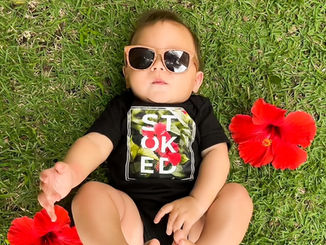 A baby laying in green grass wearing sunglasses with two hibiscus flowers next to him.  Wearing a black bodysuit that says stoked within a graphic of hibiscus flowers
