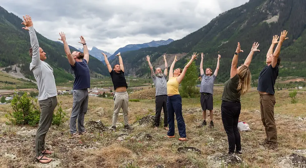 A group of hikers stretches with arms raised in a rocky alpine valley, surrounded by green mountains under a cloudy sky in a relaxed, upbeat moment.